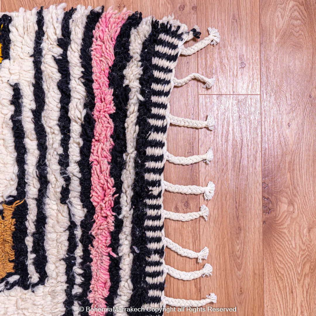 Colorful Moroccan Berber rug with pink, black, and cream stripes on a wooden floor. Handmade wool art piece.