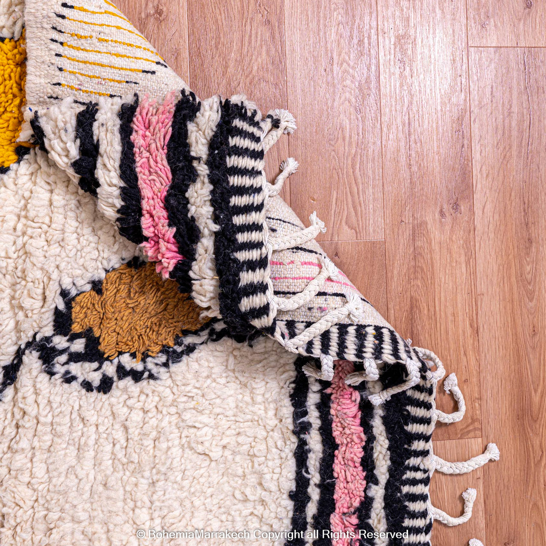 Colorful handmade Moroccan wool rug with zigzag patterns in mustard yellow, pink, black, and cream, placed on wooden flooring.