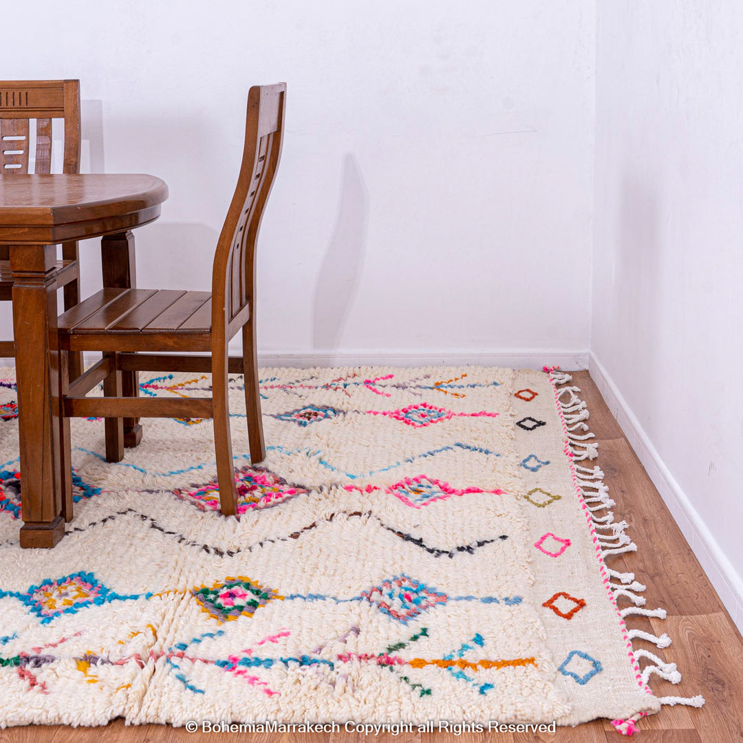 Colorful Moroccan rug with diamond Berber pattern, pink, blue, yellow on cream, under wooden dining table, enhancing room decor.