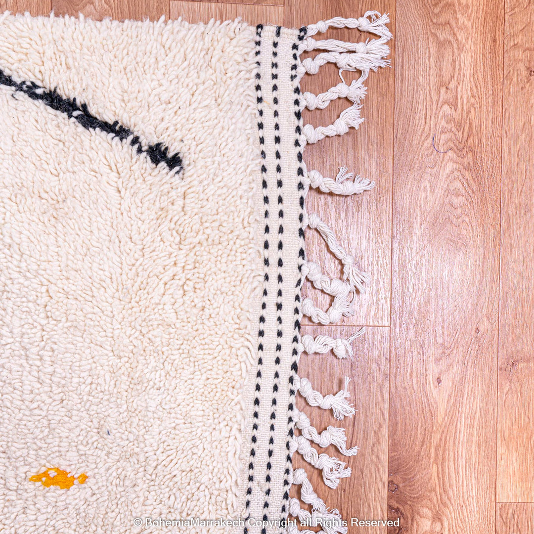 Corner of a traditional Moroccan wool rug with black and orange details, showcasing intricate weaving on a wooden floor.