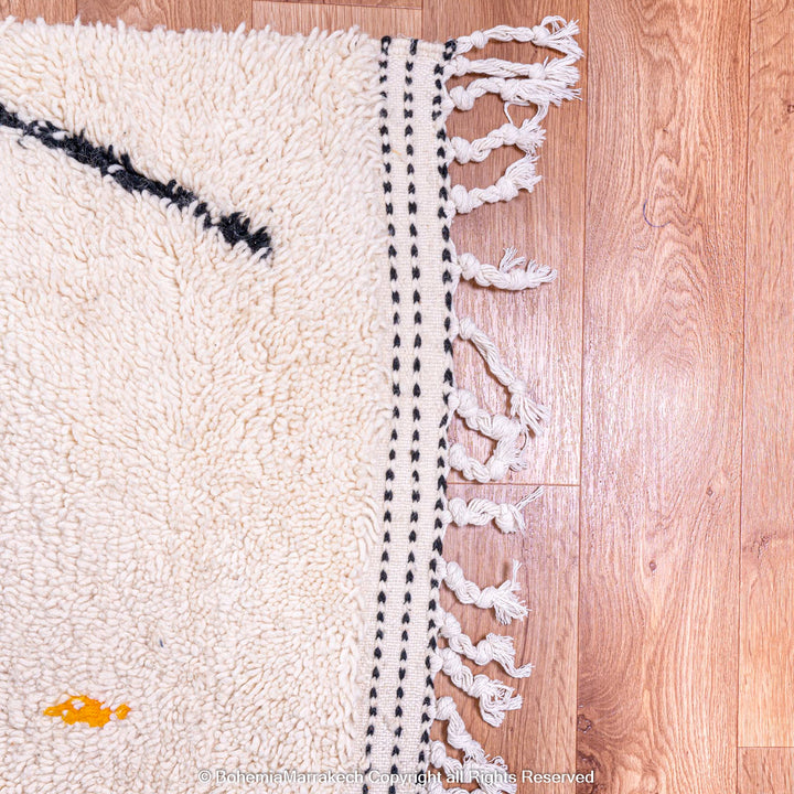 Corner of a traditional Moroccan wool rug with black and orange details, showcasing intricate weaving on a wooden floor.