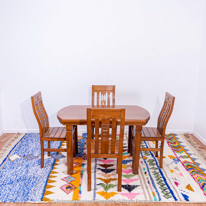 Oval wooden dining table with four chairs placed on a colorful Moroccan rug featuring geometric patterns and vibrant colors.