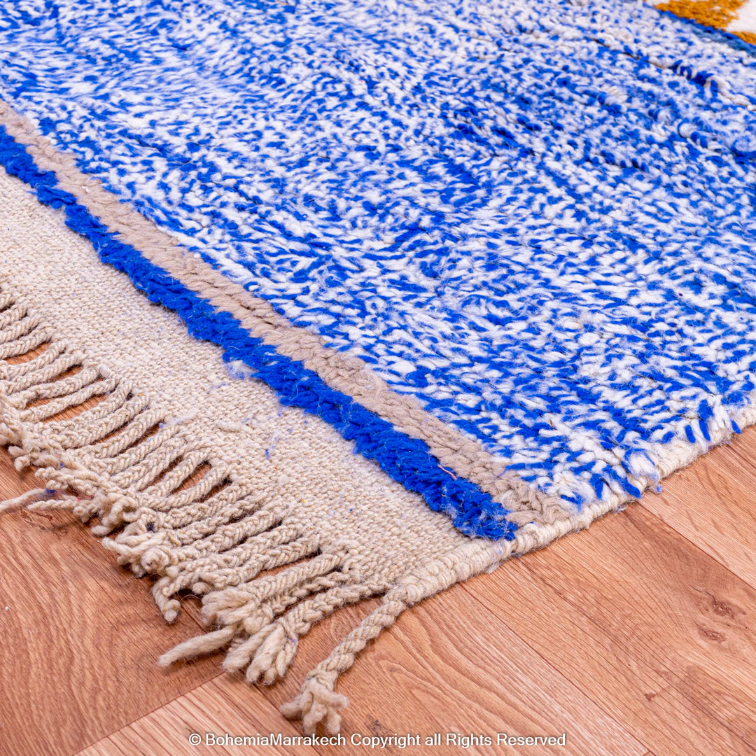 Blue and white Moroccan rug with fringe on wood floor, showcasing traditional Berber craftsmanship and geometric patterns.