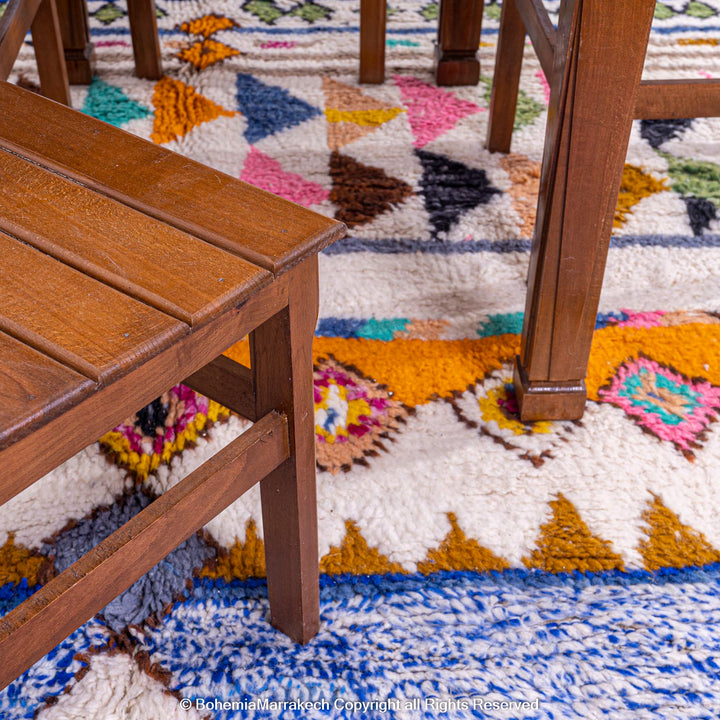 Colorful Moroccan Berber rug with geometric patterns, featuring vibrant blues, pinks, and greens, under a wooden table and chair.