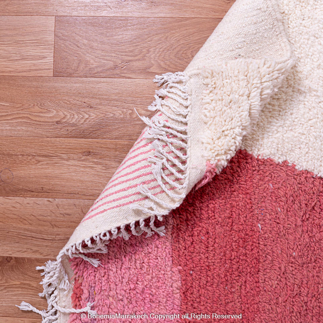 Red and pink handmade Moroccan wool rug with hourglass design and fringed edges on a wooden floor.