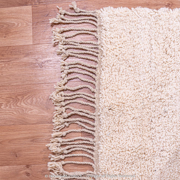 Ivory Moroccan wool rug with fringes on wood floor, traditional Beni Ourain style.