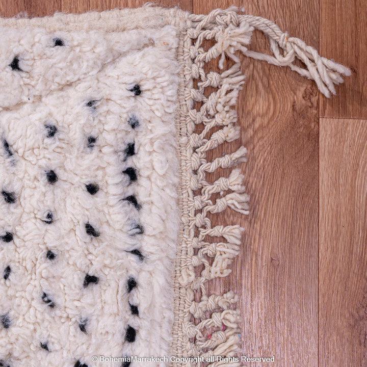 Close-up of a white Moroccan rug with black dot pattern and fringe on a wooden floor, 100% natural wool, handcrafted quality.