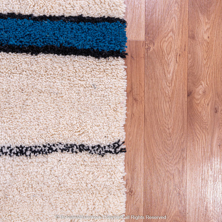 Moroccan wool rug with blue and black stripes on wooden floor, Beni Ourain style, handwoven by Berber artisans.