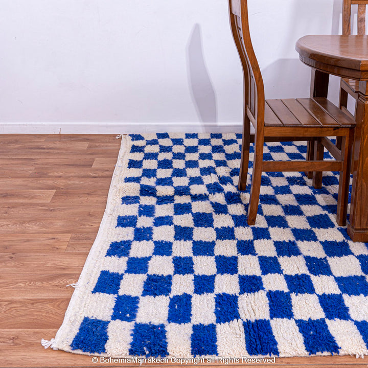 Handmade blue and white checkered Moroccan rug, wool, on wooden floor with chair, perfect for modern eclectic interior design.
