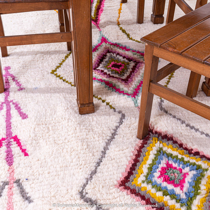 Colorful Moroccan Azilal rug with traditional diamond motifs under wooden furniture, featuring pink, blue, and yellow patterns on an ivory background.