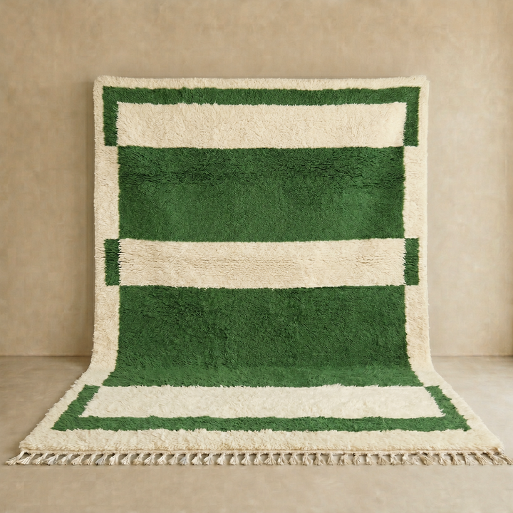 Green and cream handmade Moroccan wool rug with bold geometric border design, plush texture, and fringed tassels, displayed against a neutral beige wall.