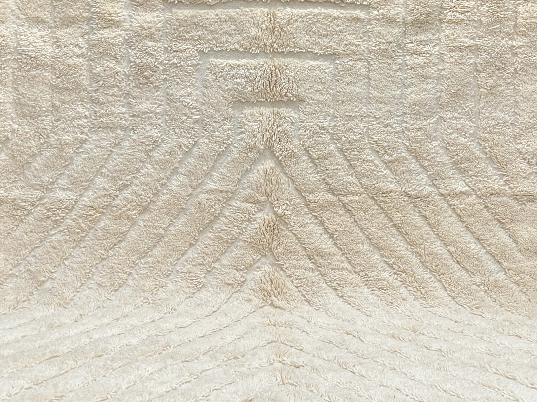 Beige textured rug with tassels on a black floor against a light gray brick wall.
