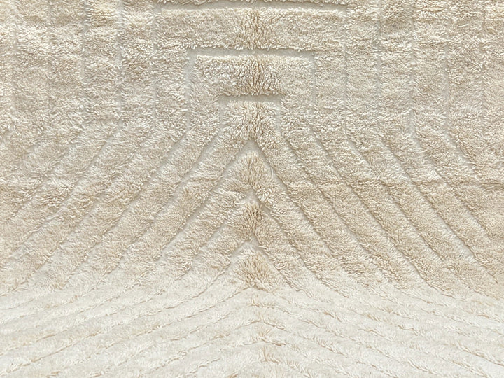 Beige textured rug with tassels on a black floor against a light gray brick wall.