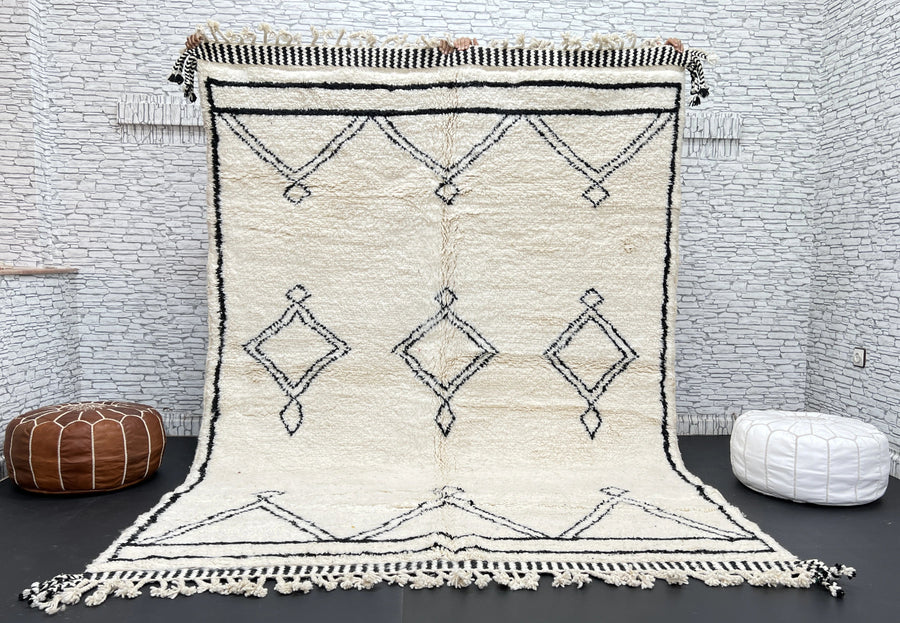 Beige and black patterned rug with tassels on a dark floor against a white brick wall.