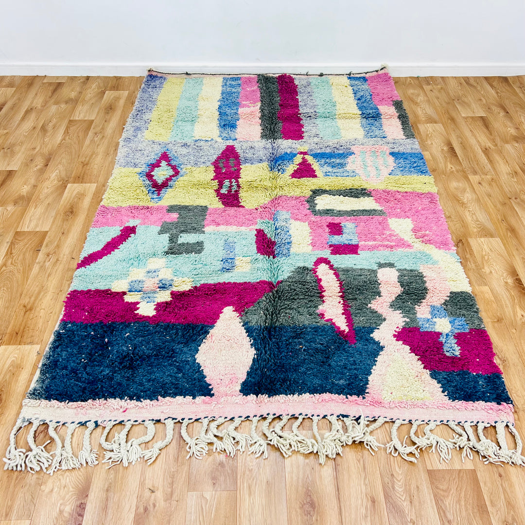 Colorful vintage Moroccan Berber rug in 8x5 ft size with pink, blue, yellow, and green geometric patterns on a wooden floor.