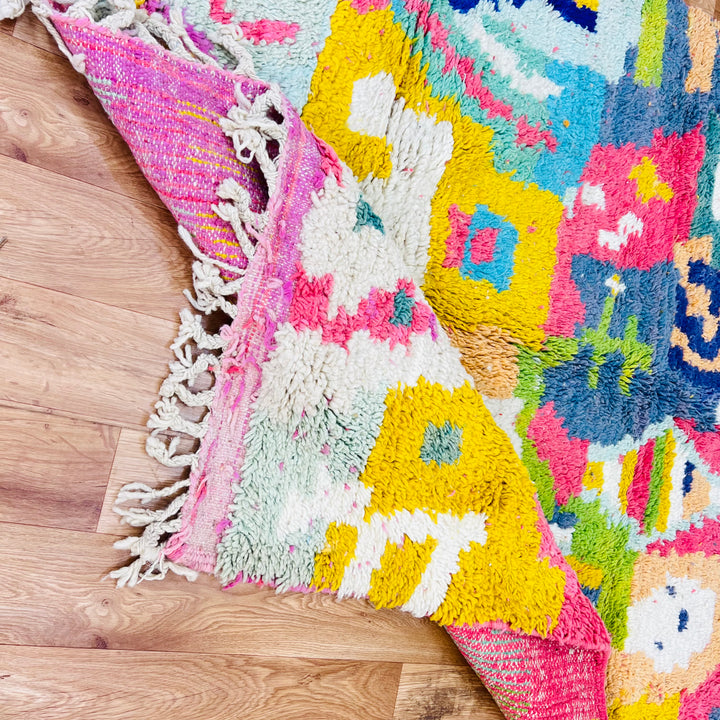 Colorful abstract Moroccan rug on wood floor, featuring bold pinks, blues, yellows, and greens, showcasing traditional Berber symbols.