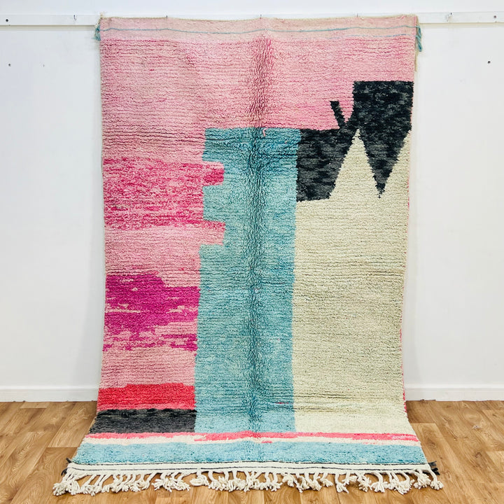 Abstract pink Moroccan rug, 8x5 ft, featuring blush pinks, aqua blues, ivory wool, and bold black shapes. Handmade Berber design.