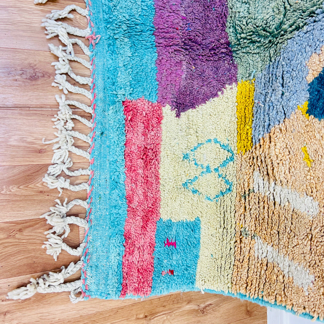 Colorful geometric Moroccan rug with abstract patterns, handwoven by Berber artisans, featuring vibrant turquoise, pink, and yellow hues.