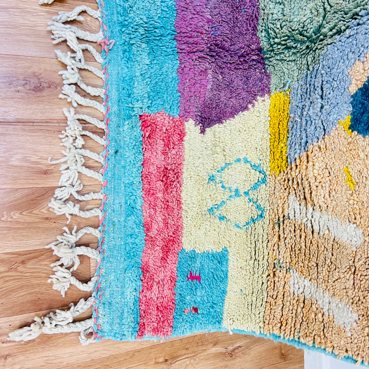 Colorful geometric Moroccan rug with abstract patterns, handwoven by Berber artisans, featuring vibrant turquoise, pink, and yellow hues.