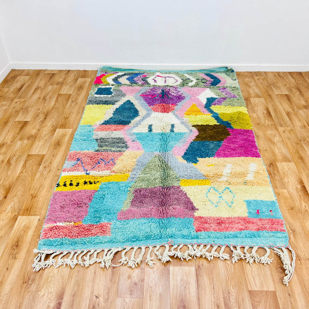 Colorful geometric Moroccan Berber wool rug in turquoise, pink, and yellow on wooden floor, 8.03 x 5.18 ft.