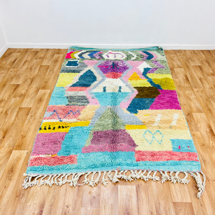Colorful geometric Moroccan Berber wool rug in turquoise, pink, and yellow on wooden floor, 8.03 x 5.18 ft.