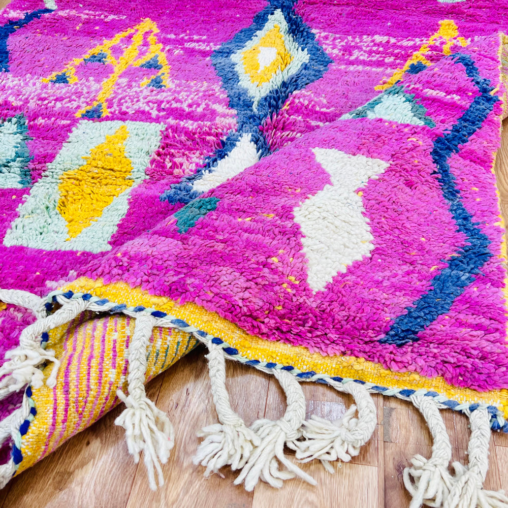 Pink Moroccan rug with tribal symbols, handwoven by Berber artisans, featuring mustard, teal, and navy motifs on wood floor.