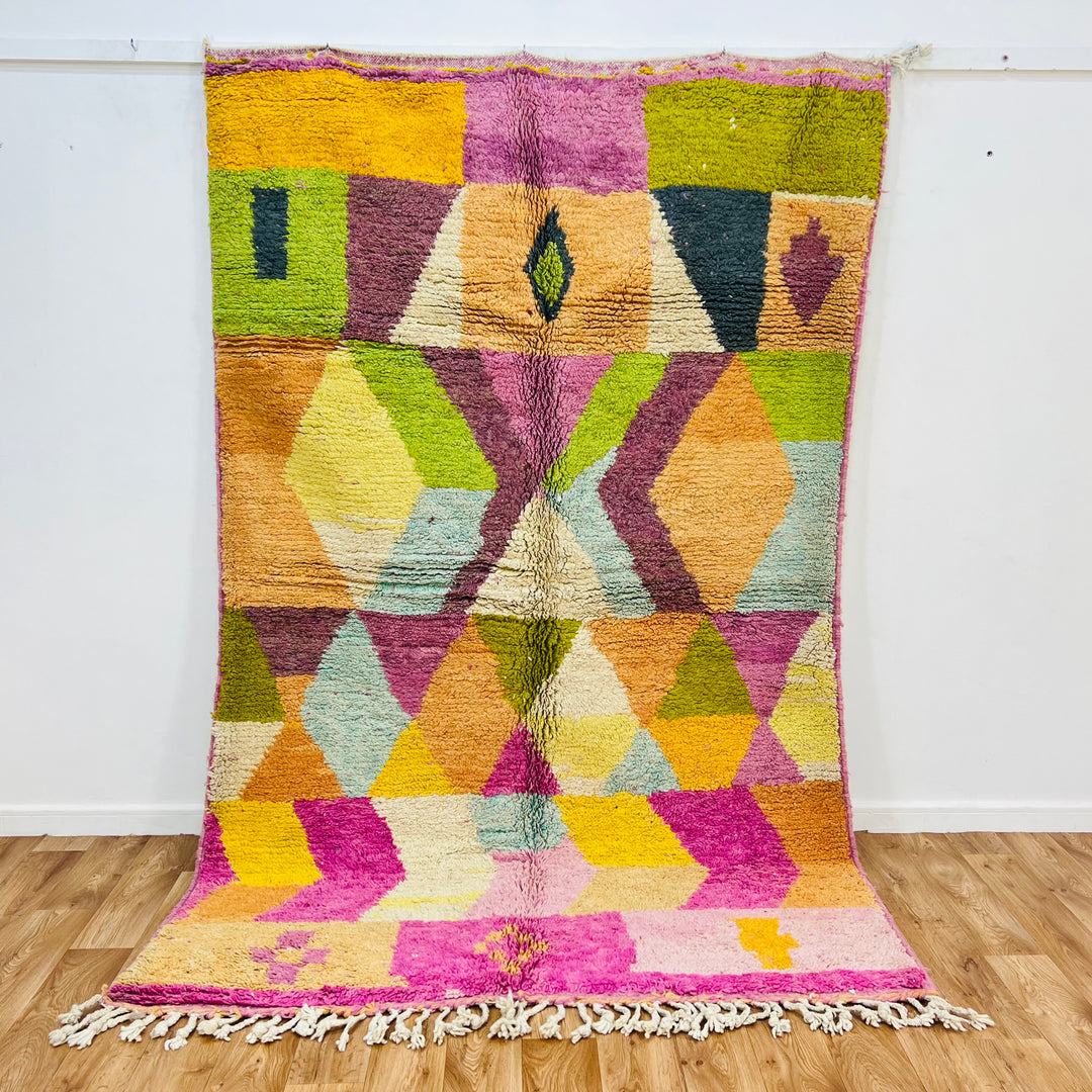 Colorful Moroccan geometric wool rug with diamond and triangle patterns in earthy tones, accented with pink tassels and a plush base.