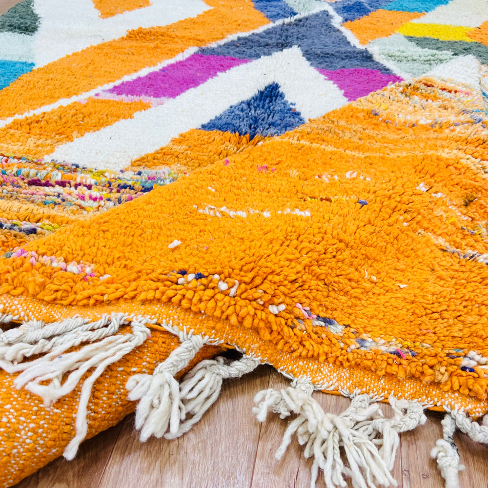 Large colorful Moroccan wool rug with bold tribal patterns in orange, navy, fuchsia, and teal, featuring fringed edges.