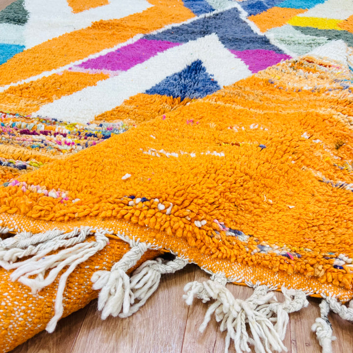 Large colorful Moroccan wool rug with bold tribal patterns in orange, navy, fuchsia, and teal, featuring fringed edges.