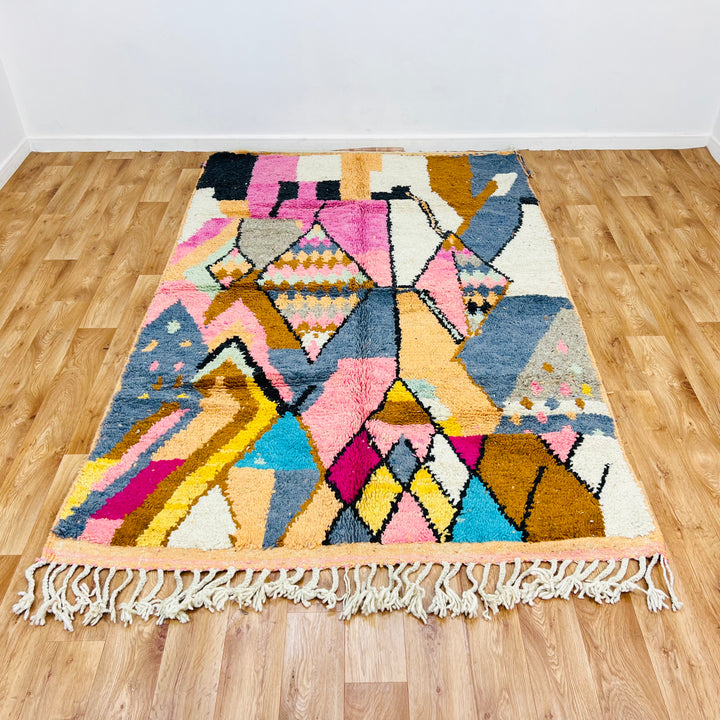 Colorful vintage Moroccan Berber wool rug with geometric diamond patterns in pink, cream, mustard, blue, and orange hues on wood floor.