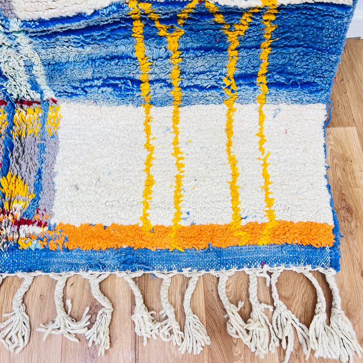Color block Moroccan Berber rug with blue, yellow, and orange patterns on wood flooring. Handwoven wool, 244x168 cm.