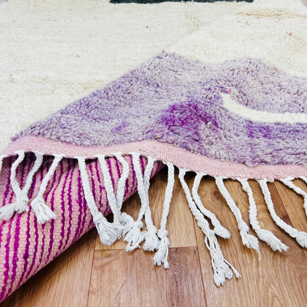 Vintage Moroccan rug with geometric design in purple and ivory tones, showing hand-knotted wool texture, on wooden floor.