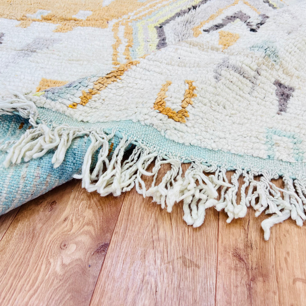 Close-up of a pastel diamond vintage Moroccan rug with fringe, showcasing intricate tribal motifs in pink, gold, and gray on wood floor.