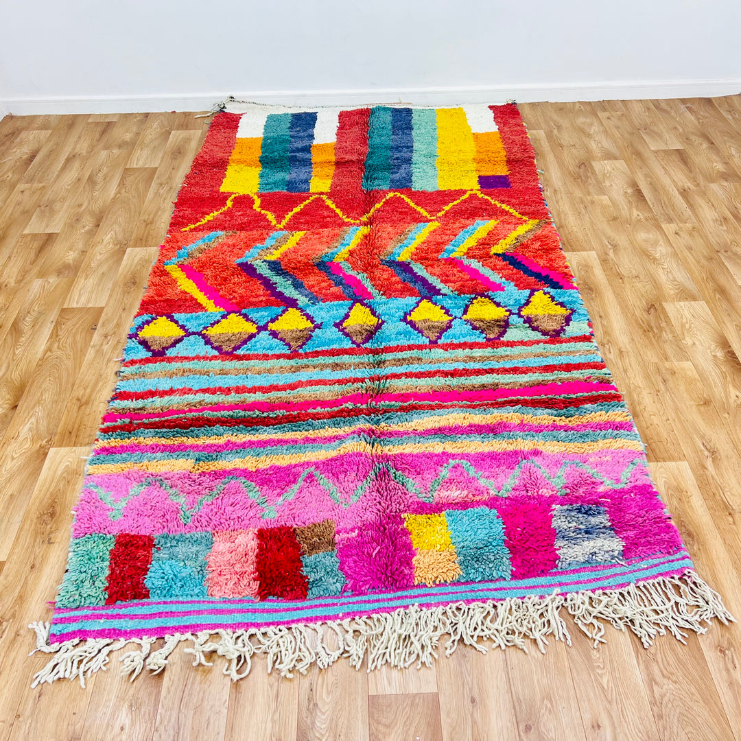 Bold colorful Moroccan Berber rug with geometric patterns in red, yellow, blue, and pink shades on wooden floor.