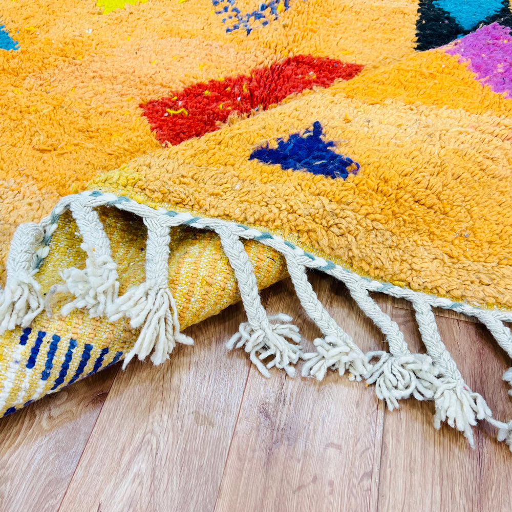 Bright orange Moroccan wool rug with tribal Berber motifs in blue, red, and pink on wooden floor, featuring tassel fringe.
