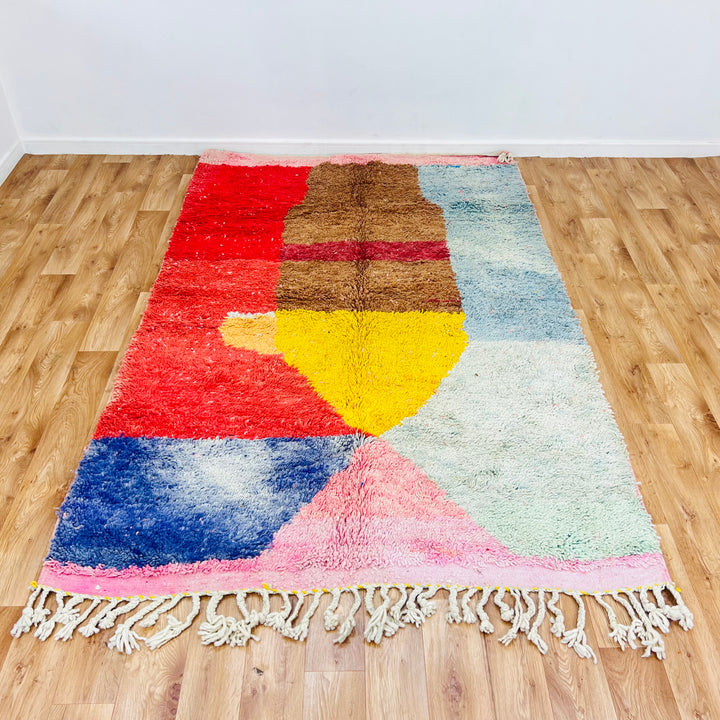 Handmade Moroccan wool rug with pink, yellow, red, and blue hues, featuring a diamond pattern on a hardwood floor.