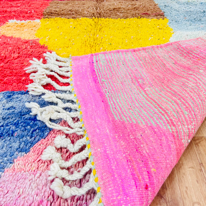 Colorful vintage Moroccan rug with pink, red, yellow, blue, and brown diamond patterns, fringed edges visible.