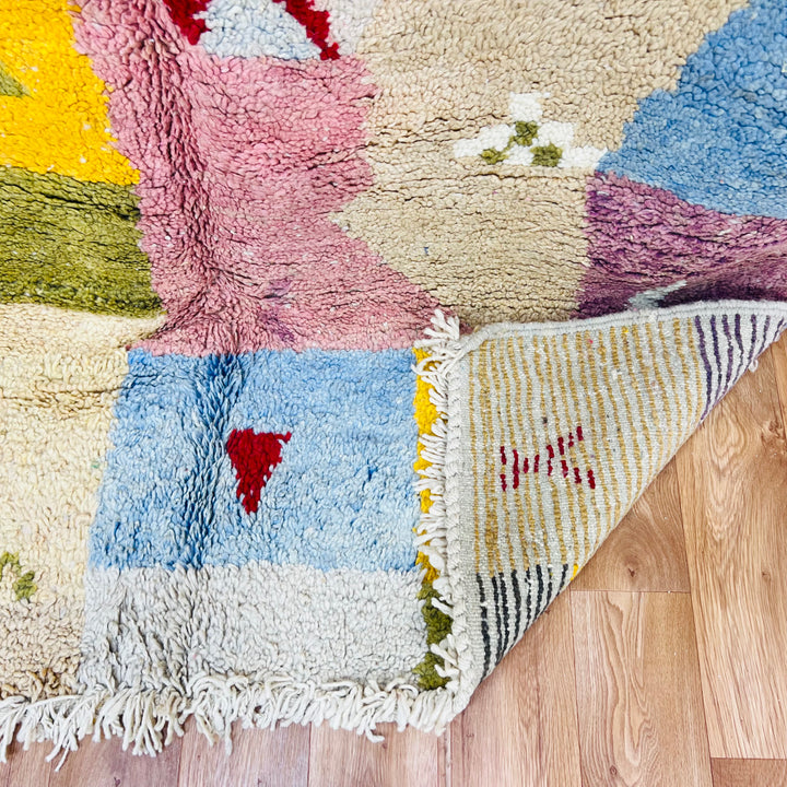 Colorful vintage Moroccan wool rug with patchwork design in pink, yellow, blue, olive green, and burgundy on wooden floor.