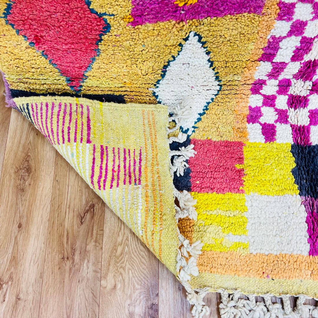 Colorful Beni Mrirt Moroccan rug with diamond and checkerboard motifs in pink, yellow, and teal on wooden floor. Authentic wool rug.