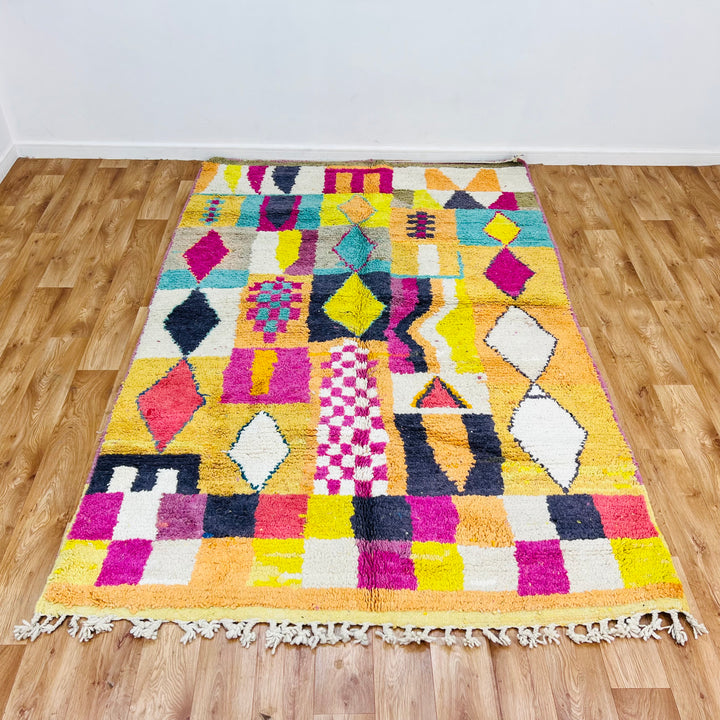 Colorful Beni Mrirt Moroccan rug with diamond and checkerboard patterns in pink, yellow, black, and teal on wooden floor.