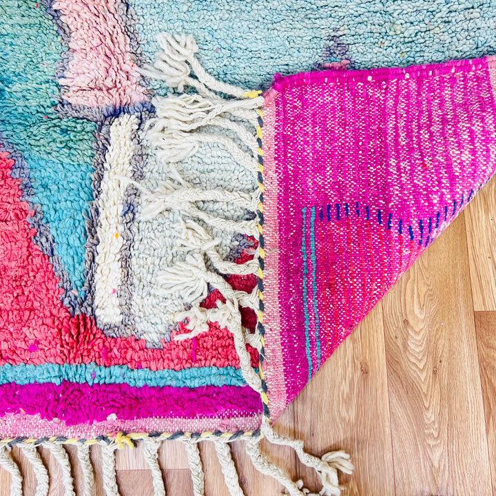 Colorful geometric Moroccan wool rug with pink, mint, and navy patterns, featuring fringe detail on the wooden floor.