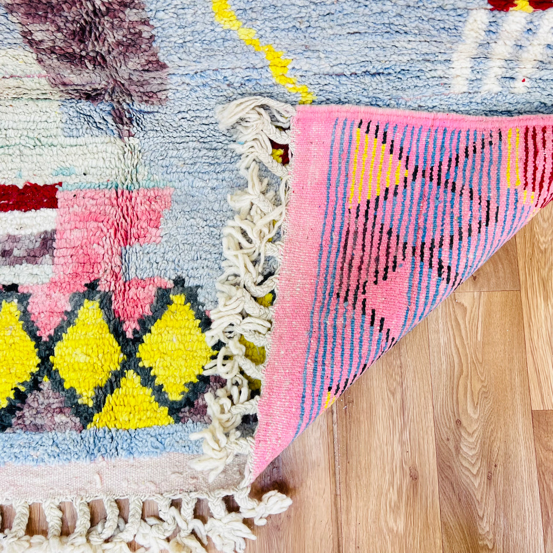 Abstract Berber Moroccan wool rug with geometric shapes in yellow, coral pink, and blue on a wooden floor.