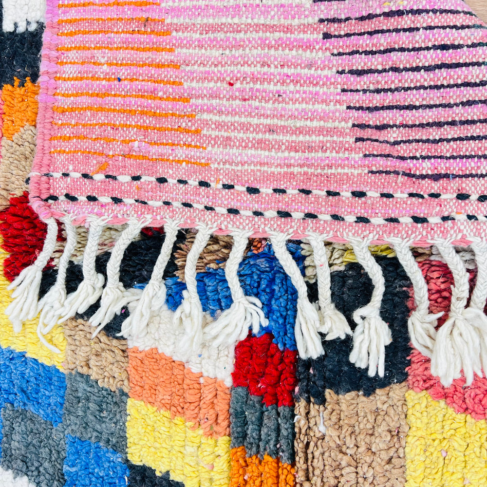 Close-up of a colorful Moroccan rug featuring geometric patterns in magenta, saffron yellow, blue, and pink with fringe detail.