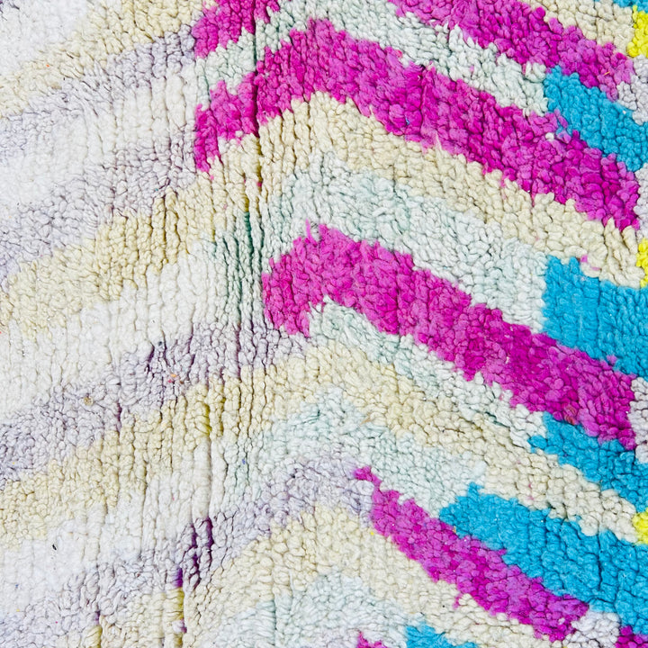 Close-up of a vintage Moroccan rug with a vibrant chevron pattern in pink, turquoise, and yellow accents on a beige background.