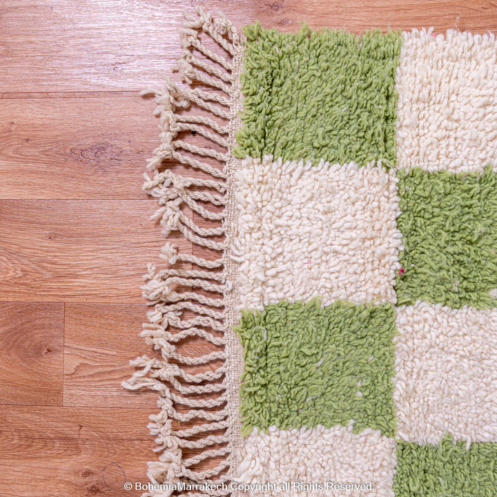 Green and white checkered Moroccan wool rug with fringe detail on wooden floor.