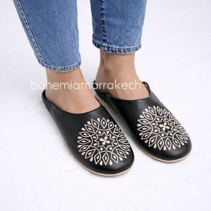 Hand-stitched black Moroccan leather babouche slippers with carved geometric design on soft yellow background