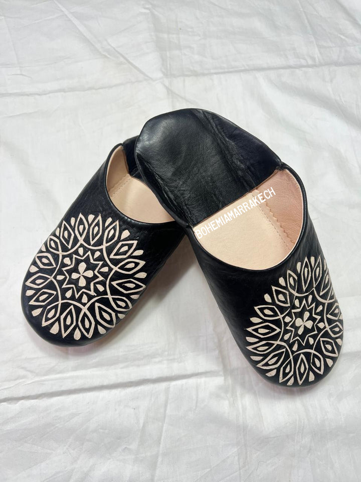 Hand-stitched black Moroccan leather babouche slippers with carved geometric design on soft yellow background