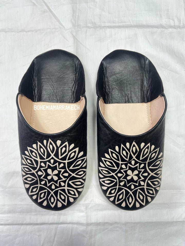 Hand-stitched black Moroccan leather babouche slippers with carved geometric design on soft yellow background