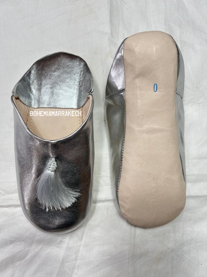 Silver Moroccan babouche slippers with tassel detail on light yellow background