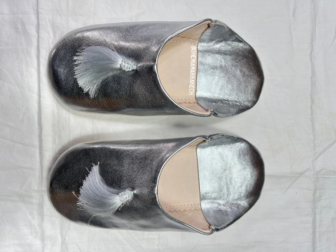 Silver Moroccan babouche slippers with tassel detail on light yellow background
